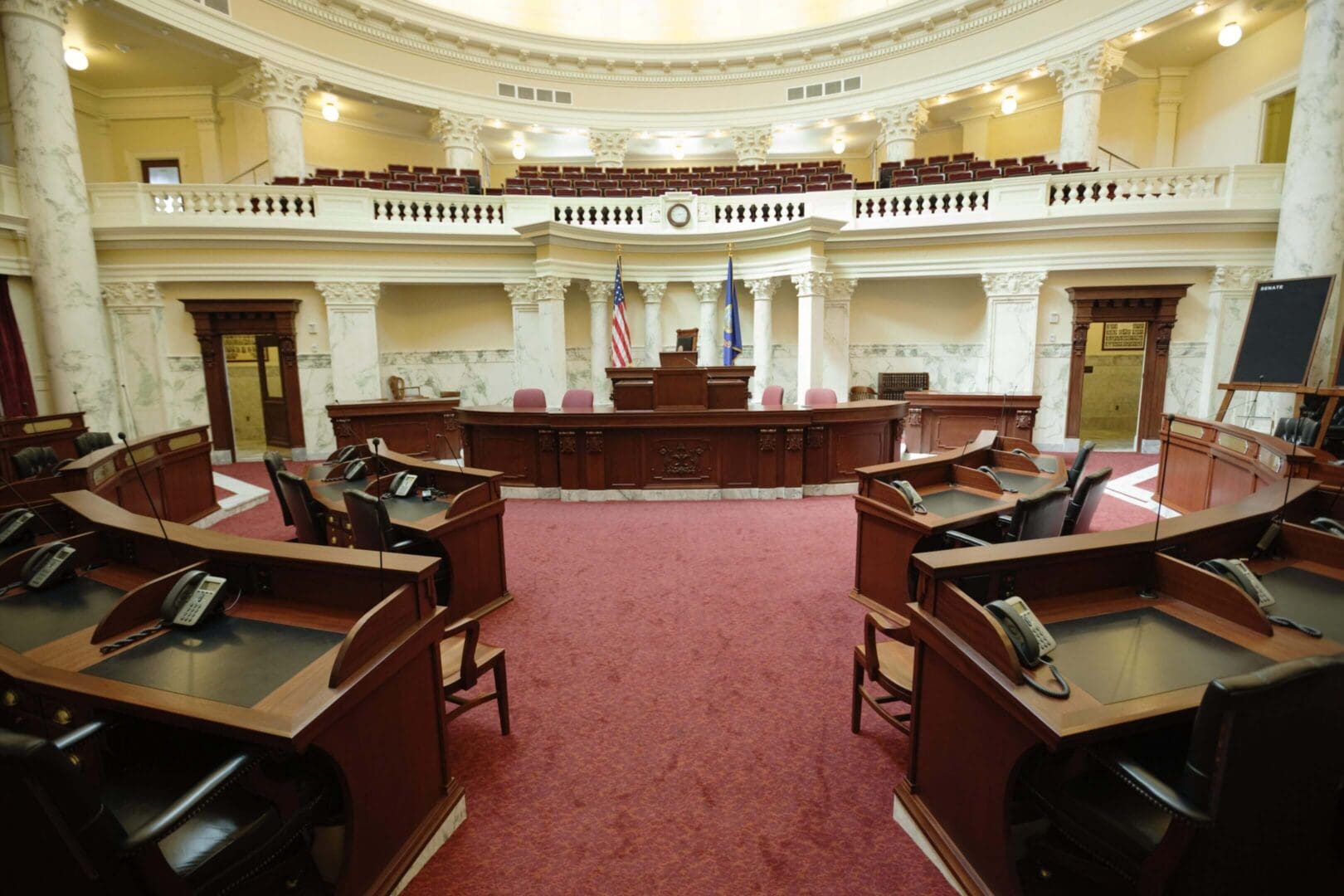 Empty legislative chamber with wooden desks and red carpet.